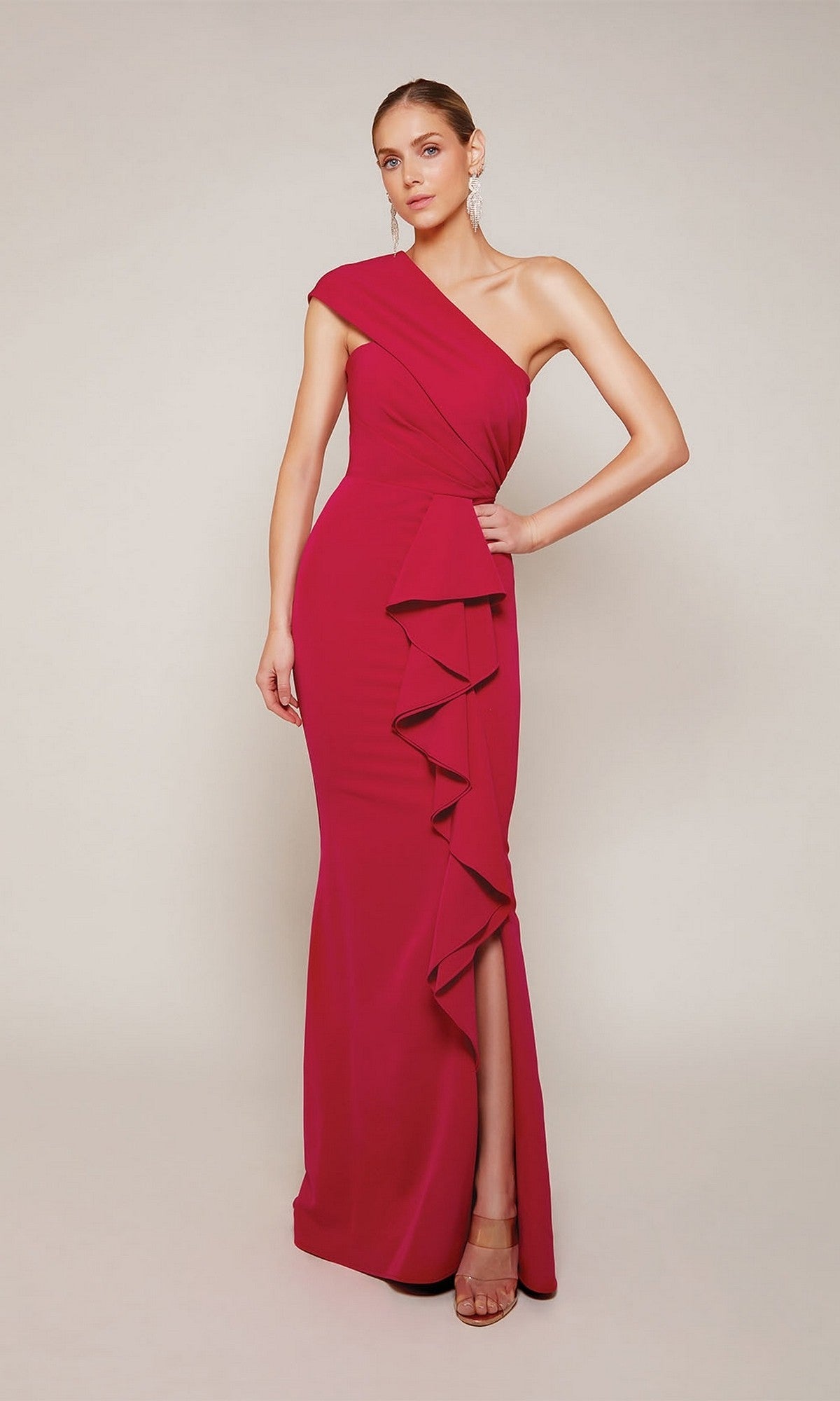 A woman wearing the Alyce 27646 long formal dress in raspberry, featuring a one-shoulder design, ruffled detail, sheath silhouette, and thigh-high slit, stands against a plain light-colored background.